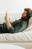 Ein Mann liegt auf einem Sofa und trägt Kopfhörer, während er sein Smartphone bedient.